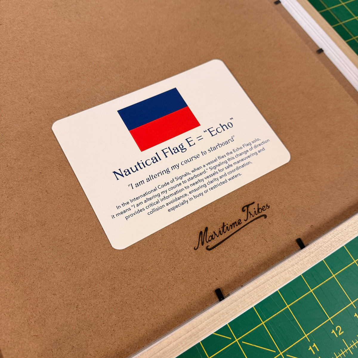 A close-up of a wooden framed print or plaque on brown kraft backing, showing a white label with a Russian flag graphic and the text 'Nautical Flag E = "Echo"' plus a script brand name 'Maritime Tides.' A green cutting mat is visible around the frame.