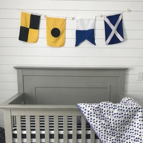 A gray wooden crib with a white and navy blue patterned blanket draped over the side, set in a nursery with a white shiplap wall, nautical flag bunting, and a white fluffy rug.